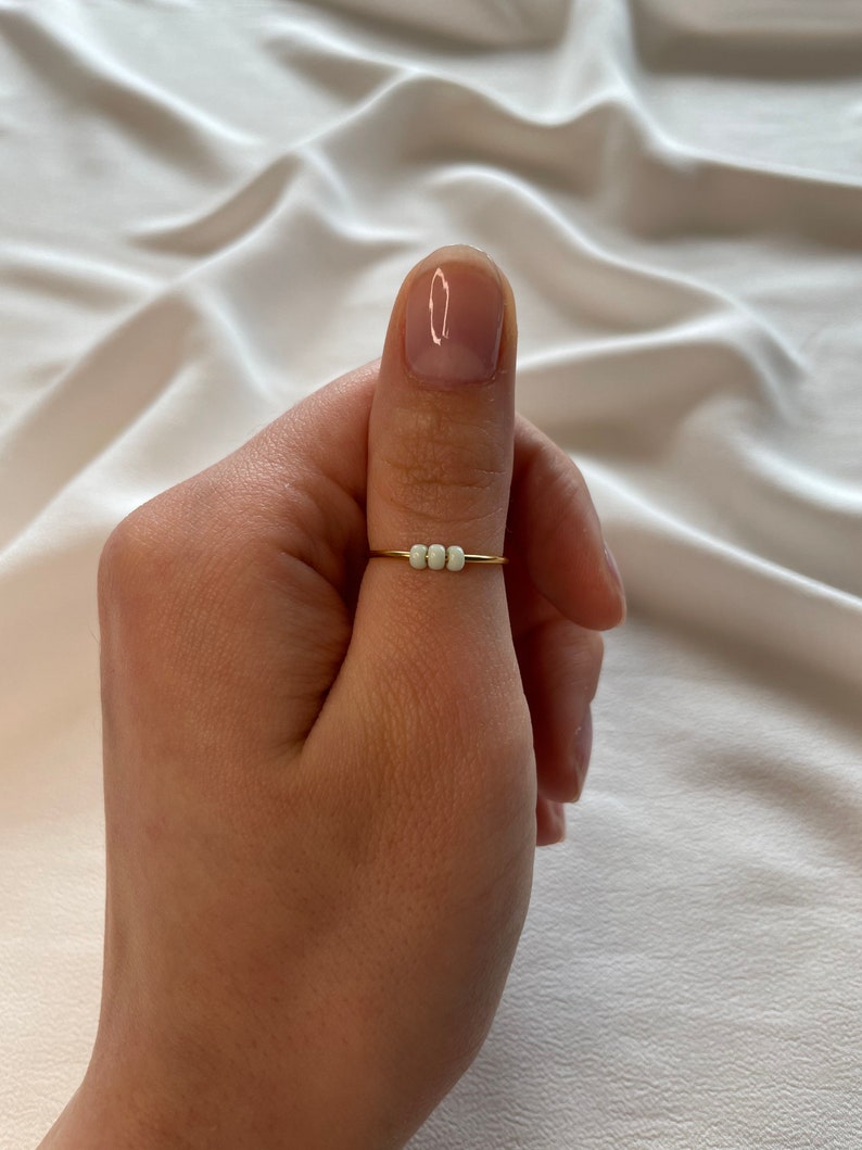 May include: A gold ring with three white beads on a person's thumb.