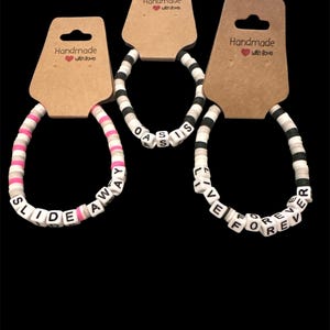 May include: Three handmade bracelets with white letter beads spelling "SLIDE AWAY", "OASIS", and "LIVE FOREVER". The bracelets have colorful disc beads in pink, black, white, and gray. Each bracelet is attached to a brown tag that says "Handmade with love".