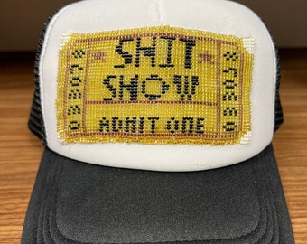 Op de afbeelding: Witte truckercap met een zwart mesh achterpaneel. De voorkant van de cap heeft een geel en zwart geperst patch met de tekst "SHIT SHOW" met daaronder een kleiner geperst patch met de tekst "ANYTIME".