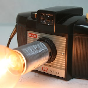 Puede incluir: Una cámara Kodak 127 negra vintage con una lente plateada y una bombilla brillante adjunta. La cámara tiene un logotipo Kodak rojo y blanco y un dial color crema en la parte superior. Un equipo de fotografía clásico.
