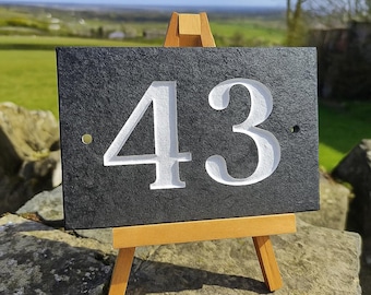 May include: A black slate house number sign with the number 43 carved in white. The sign is displayed on a small wooden easel, set against a backdrop of a green field and blue sky.