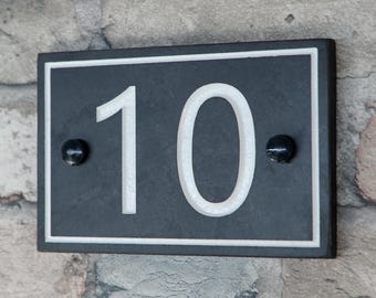 May include: Rectangular black house number plaque with the number "10" in white. The number is centered on the plaque, which has a white border. Two black, round accents are on either side of the number. The plaque is mounted on a stone wall.