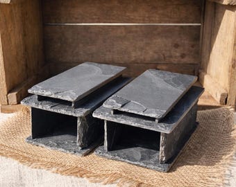 May include: Two rectangular, dark gray slate serving trays with a rustic, natural texture. Each tray has a raised base, creating a tiered design. The trays are displayed on a burlap surface against a wooden background.
