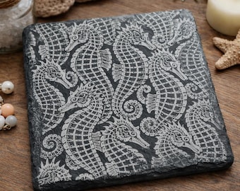 May include: Square, dark gray slate coaster with a white seahorse pattern. The seahorses are intricately detailed, creating a repeating design across the coaster's surface. The coaster is likely used for drinks and adds a decorative touch to any space.