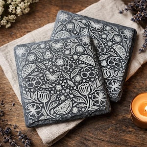 Pode incluir: Duas bases quadradas de ardósia com um design floral e frutado branco, incluindo borboletas, cerejas e frutas cítricas. As bases repousam sobre um pano de linho bege, com um fundo de madeira e uma vela acesa.