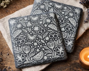 May include: Two square slate coasters with a white floral and fruit design, including butterflies, cherries, and citrus. The coasters rest on a beige linen cloth, with a wooden background and a lit candle in a small copper-colored holder.
