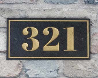 May include: A rectangular black house number plaque with gold-colored numbers and trim. The numbers "321" are prominently displayed in a bold, serif font. The plaque is mounted on a multi-colored brick wall.
