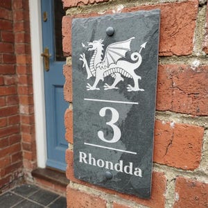 May include: A rectangular dark grey slate house sign featuring a white engraved Welsh dragon at the top, the number 3 in the centre, and the word Rhondda at the bottom. The sign is secured with two metal bolts at the top and bottom centre.