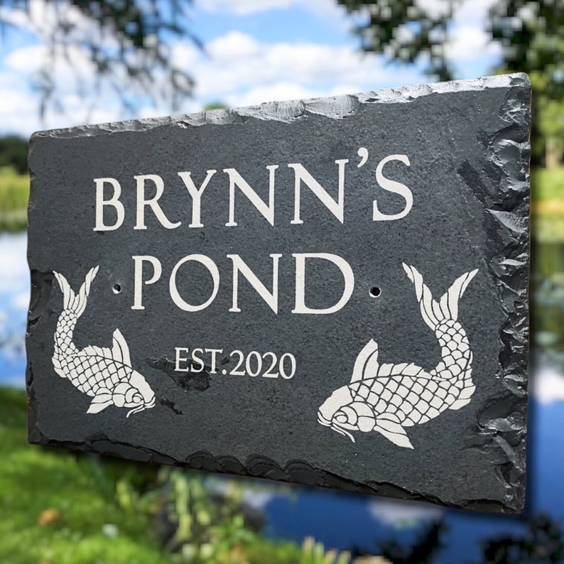 May include: A rectangular black slate sign with rough, chiseled edges. The text reads BRYNN'S POND and EST. 2020 in white lettering. Two white engraved koi fish illustrations are positioned on the left and right sides of the word POND.