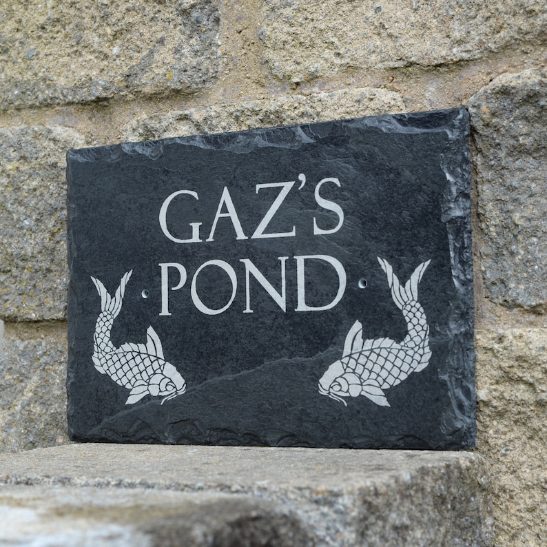 May include: A rectangular black slate sign with rough, natural edges. The text GAZ'S POND is engraved in the centre in a light grey font. Two detailed fish illustrations are etched on either side of the word POND, and two small holes are visible.