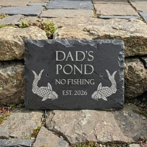 May include: A rectangular dark grey slate sign with rough, uneven edges. The text DAD'S POND NO FISHING EST. 2026 is engraved in white. Two stylised fish illustrations are positioned on the left and right sides of the text, facing inward.