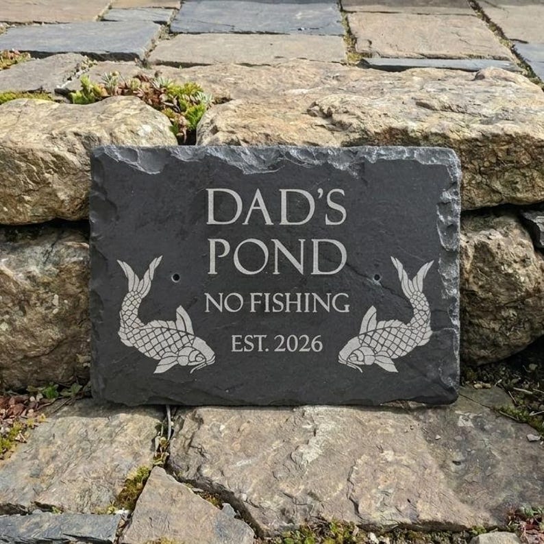 May include: A rectangular dark grey slate sign with rough, uneven edges. The text DAD'S POND NO FISHING EST. 2026 is engraved in white. Two stylised fish illustrations are positioned on the left and right sides of the text, facing inward.