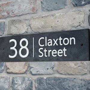 May include: A rectangular black slate house number sign with the number "38" and the words "Claxton Street" in white lettering. The sign is mounted on a multi-colored brick wall.