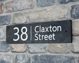 May include: A rectangular black slate house number sign with the number "38" and the words "Claxton Street" in white lettering. The sign is mounted on a multi-colored brick wall.