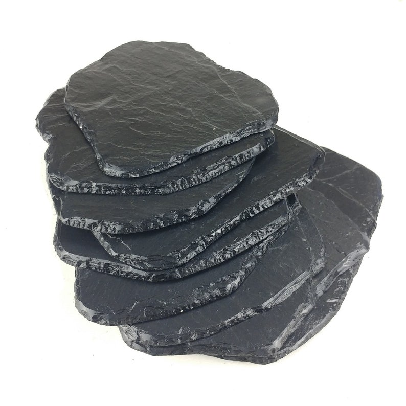 May include: A stack of irregularly shaped, black slate coasters. The coasters have rough, textured edges and a dark, natural color. They are arranged in a slightly fanned-out pattern, showcasing their unique shapes and surfaces.