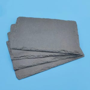 May include: A stack of four rectangular slate boards with dark grey, textured surfaces and natural, rough-hewn edges. Each board features a flat, matte finish and a distinctively uneven, layered stone texture along the perimeter of the rectangular shape.