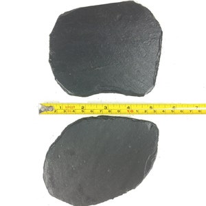 May include: Two irregularly shaped, dark gray slate serving platters. A yellow measuring tape shows the length of the platters. The platters have a rough, textured surface and are placed on a white background.