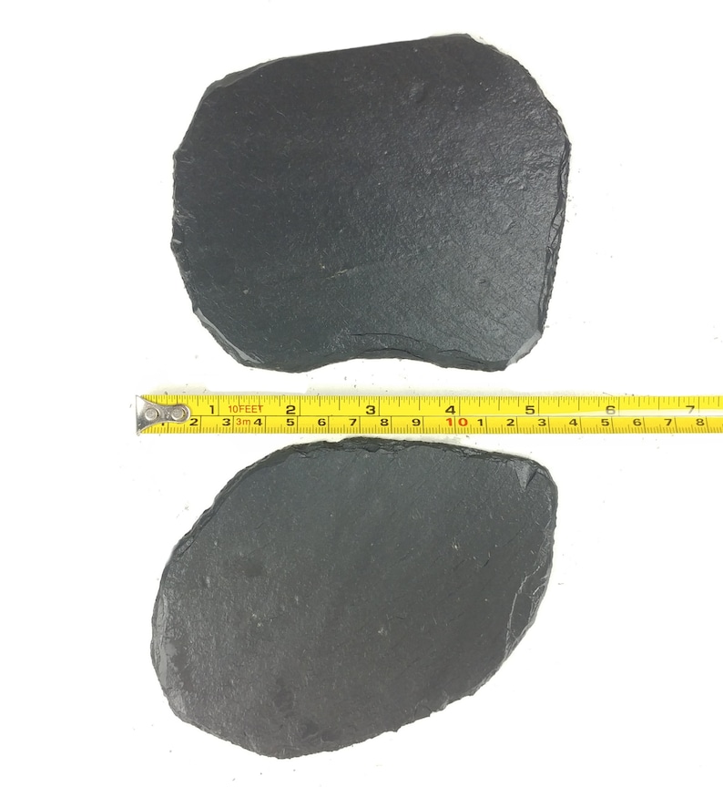 May include: Two irregularly shaped, dark gray slate serving platters. A yellow measuring tape shows the length of the platters. The platters have a rough, textured surface and are placed on a white background.