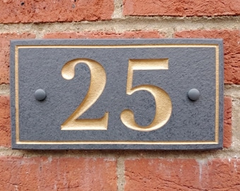 May include: A rectangular house number plaque featuring the number 25. The plaque has a dark gray, textured surface with a gold-colored border and gold-colored numerals. Two dark gray circular fasteners are positioned on the left and right sides.