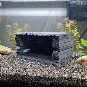 May include: A rectangular aquarium cave constructed from stacked dark grey slate stone slabs. The structure features a hollow interior space with a wide front opening, providing a shelter for small neon tetra fish swimming nearby.