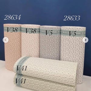 Puede incluir: Varios rollos de tela con estampado floral en varios colores, incluyendo rosa, gris y blanco. Los rollos de tela están etiquetados con números y letras, y el texto "WELCOME HOME" es visible en las etiquetas. La imagen también incluye los números "28634" y "28633".