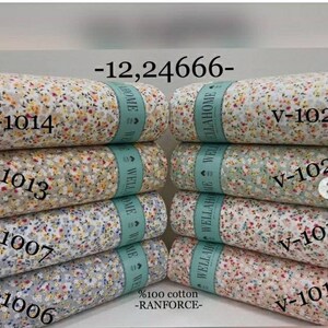 Puede incluir: Una pila de tela blanca con estampado floral. La tela está etiquetada con el texto "WELLAHOME" y el código "V-1014". La tela es 100% algodón y está etiquetada "RANFORCE".