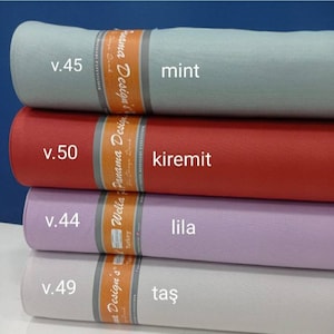 Op de afbeelding: Vier rollen stof in verschillende kleuren, gelabeld met hun namen en versienummers. De kleuren zijn mint, kiremit, lila en taş.