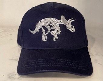 May include: A navy blue baseball cap with a white embroidered skeleton triceratops design.