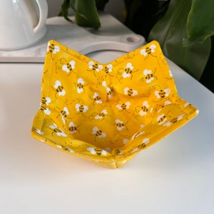 May include: Yellow fabric bowl with a bee pattern. The bowl is folded and ready to be used.