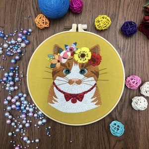 May include: A yellow embroidery hoop with a brown and white cat embroidered on it. The cat is wearing a red bow tie and a flower crown. The background is a wooden surface.