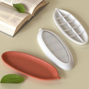 May include: Three leaf-shaped trays in white and coral colors. One white tray has a grid pattern, while the others have a leaf vein design. A green leaf and an open book are also present.