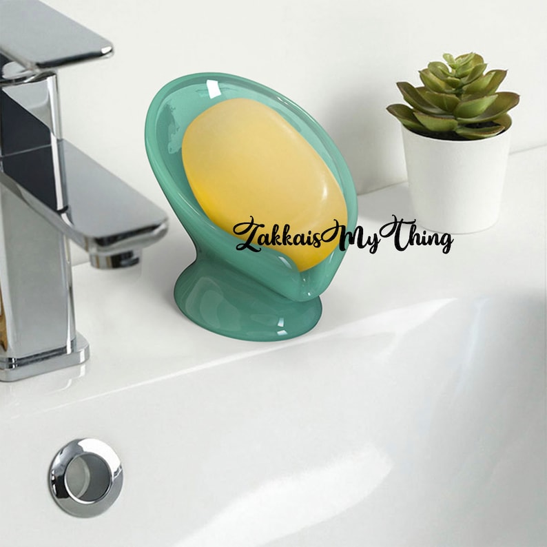 May include: A teal soap dish with a yellow bar of soap. The dish has a curved design and a pedestal base. The text "Zakkais My Thing" is written on the right side of the soap dish. A small potted plant is in the background.