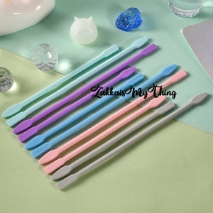 May include: A set of ten silicone stirring sticks in various pastel colors, including light blue, purple, pink, and gray. The sticks are arranged in a row, with the tips of the sticks pointing to the right. The sticks are all the same size and shape, and they have a rounded tip.