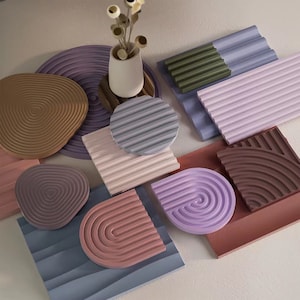 Puede incluir: Un conjunto de nueve posavasos decorativos en varios colores y patrones. Los posavasos están hechos de un material texturizado y presentan diseños geométricos. Los posavasos están dispuestos en un patrón circular sobre una superficie gris claro.