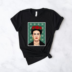 Puede incluir: Camiseta negra con un estampado gr&aacute;fico de Frida Kahlo con una corona de flores. El fondo es verde con un patr&oacute;n geom&eacute;trico. El texto "Casas Chingonas" est&aacute; impreso en la parte inferior de la camiseta.