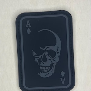 May include: Black patch in the shape of a playing card, featuring a skull design. The card shows the Ace of Spades, with the letter "A" at the top and bottom. The skull is in a lighter shade of grey.