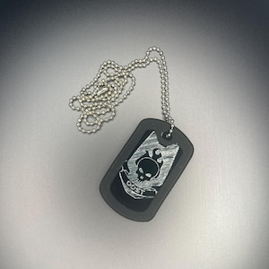 May include: Black dog tag pendant with a silver ball chain. The tag features a skull design with the text "ODST" in a silver color. The pendant is rectangular with rounded corners.