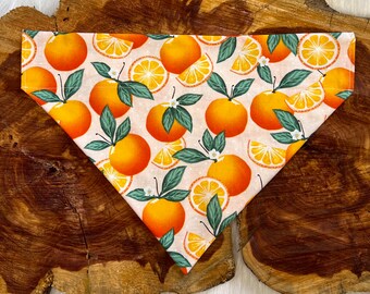 May include: Triangular dog bandana with a repeating pattern of oranges, orange slices, green leaves, and white blossoms on a light beige background. The bandana is made of fabric and is displayed on a wooden surface.