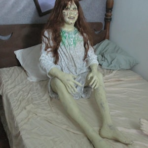 May include: A life-sized doll with long brown hair, pale green skin, and a distressed facial expression. The doll wears a white long-sleeved nightgown with a green stain on the chest, featuring a button-up collar and a patterned fabric design.