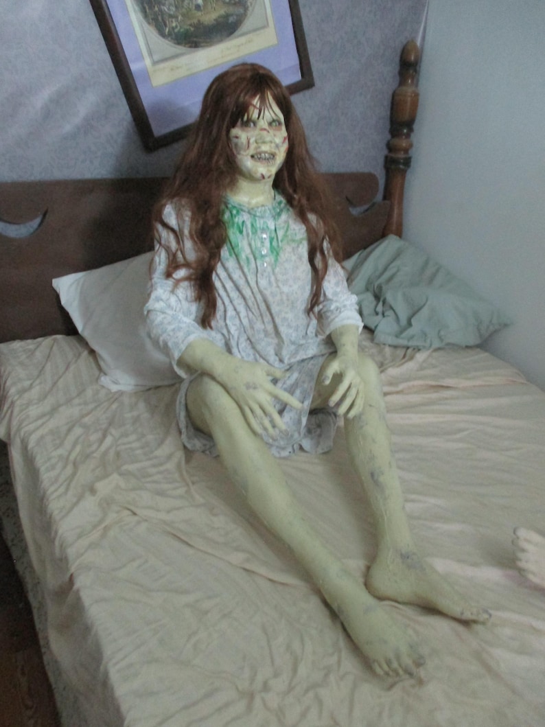 May include: A life-sized doll with long brown hair, pale green skin, and a distressed facial expression. The doll wears a white long-sleeved nightgown with a green stain on the chest, featuring a button-up collar and a patterned fabric design.