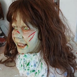 May include: A creepy Halloween prop featuring a pale-skinned face with green eyes and red slash marks. The prop has brown hair and a white collar with a floral pattern. The mouth is open, revealing teeth.
