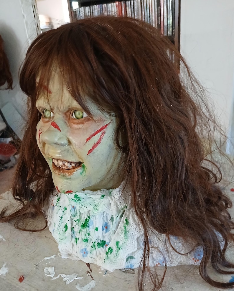 May include: A creepy Halloween prop featuring a pale-skinned face with green eyes and red slash marks. The prop has brown hair and a white collar with a floral pattern. The mouth is open, revealing teeth.