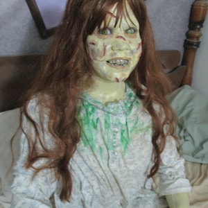 May include: A life-sized doll with long auburn hair and pale, greenish skin featuring red markings. The doll has bright green eyes, a wide grin with visible teeth, and wears a white long-sleeved nightgown with a blue floral pattern and green stains.