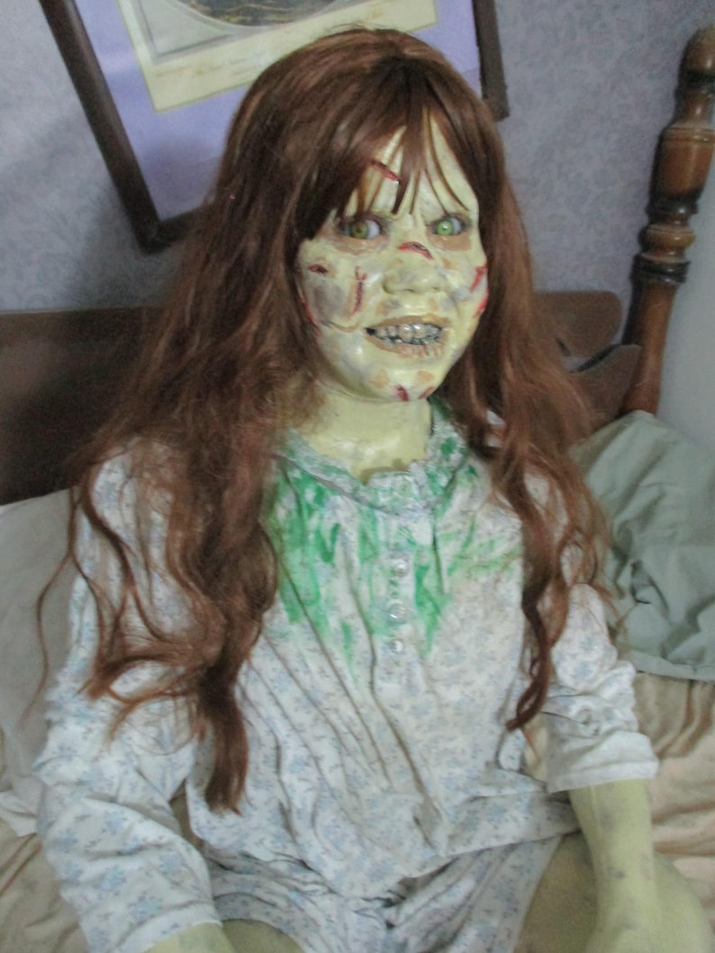 May include: A life-sized doll with long auburn hair and pale, greenish skin featuring red markings. The doll has bright green eyes, a wide grin with visible teeth, and wears a white long-sleeved nightgown with a blue floral pattern and green stains.