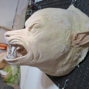 May include: A close-up of a werewolf mask made of plaster or clay. The mask is painted a light beige color and has a snarling expression with sharp teeth. The mask is mounted on a pegboard.