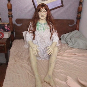 May include: A life-sized figure of a person with long auburn hair, pale skin, and a distressed facial expression. The figure wears a white nightgown with a subtle floral pattern and green stains around the neckline, sitting upright on a bed.