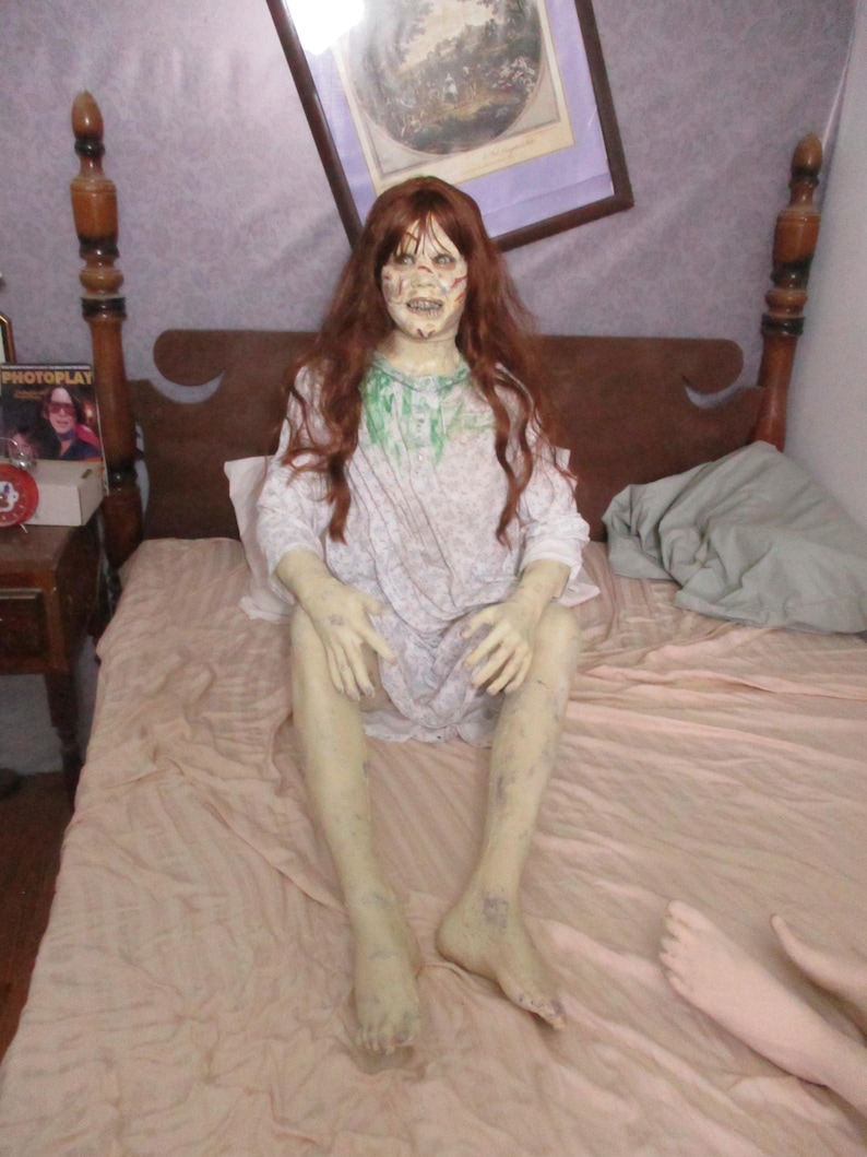 May include: A life-sized figure of a person with long auburn hair, pale skin, and a distressed facial expression. The figure wears a white nightgown with a subtle floral pattern and green stains around the neckline, sitting upright on a bed.