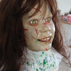 May include: A detailed, unsettling Halloween prop of a demonic girl's head. The face is pale with green eyes, red cuts, and a disturbing grin. The brown hair is long and messy, and the neck is adorned with a white floral-patterned collar.