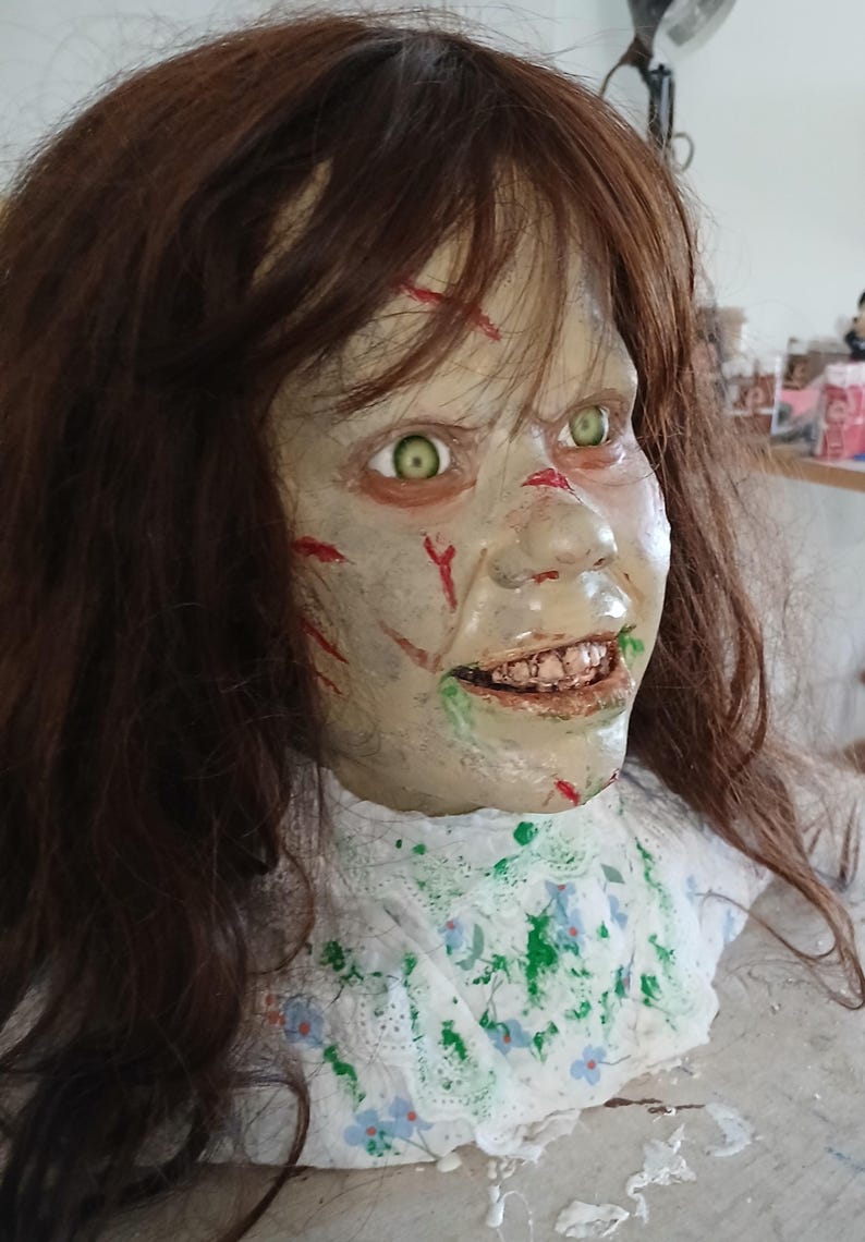 May include: A detailed, unsettling Halloween prop of a demonic girl's head. The face is pale with green eyes, red cuts, and a disturbing grin. The brown hair is long and messy, and the neck is adorned with a white floral-patterned collar.