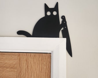 May include: Black silhouette of a cat holding a knife, mounted on a white wall above a wooden door.